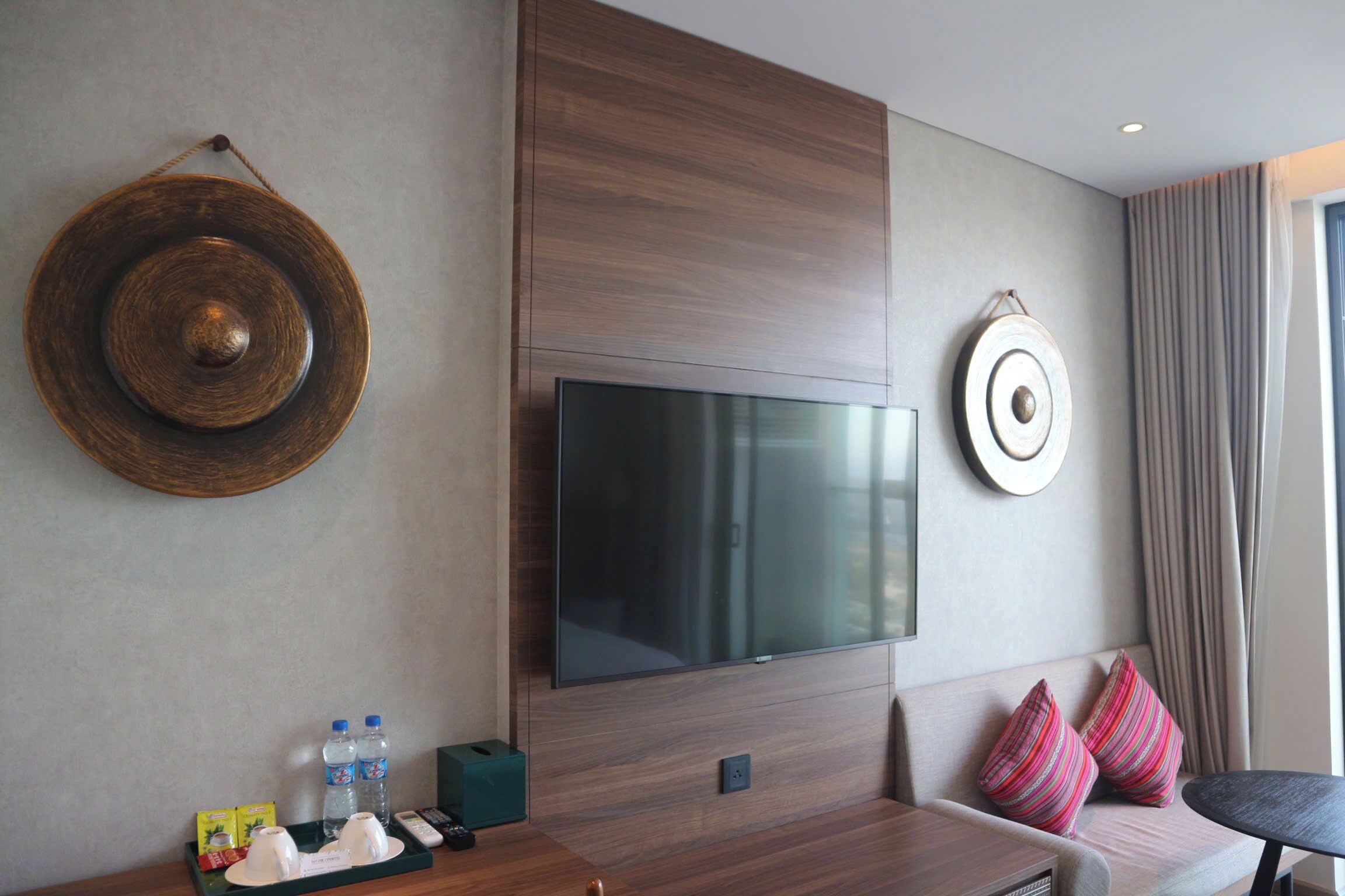 A modern hotel room with a large flat-screen TV mounted on the wall, a wooden console table with a tray of amenities, a sofa with striped pillows, and a large window with curtains.