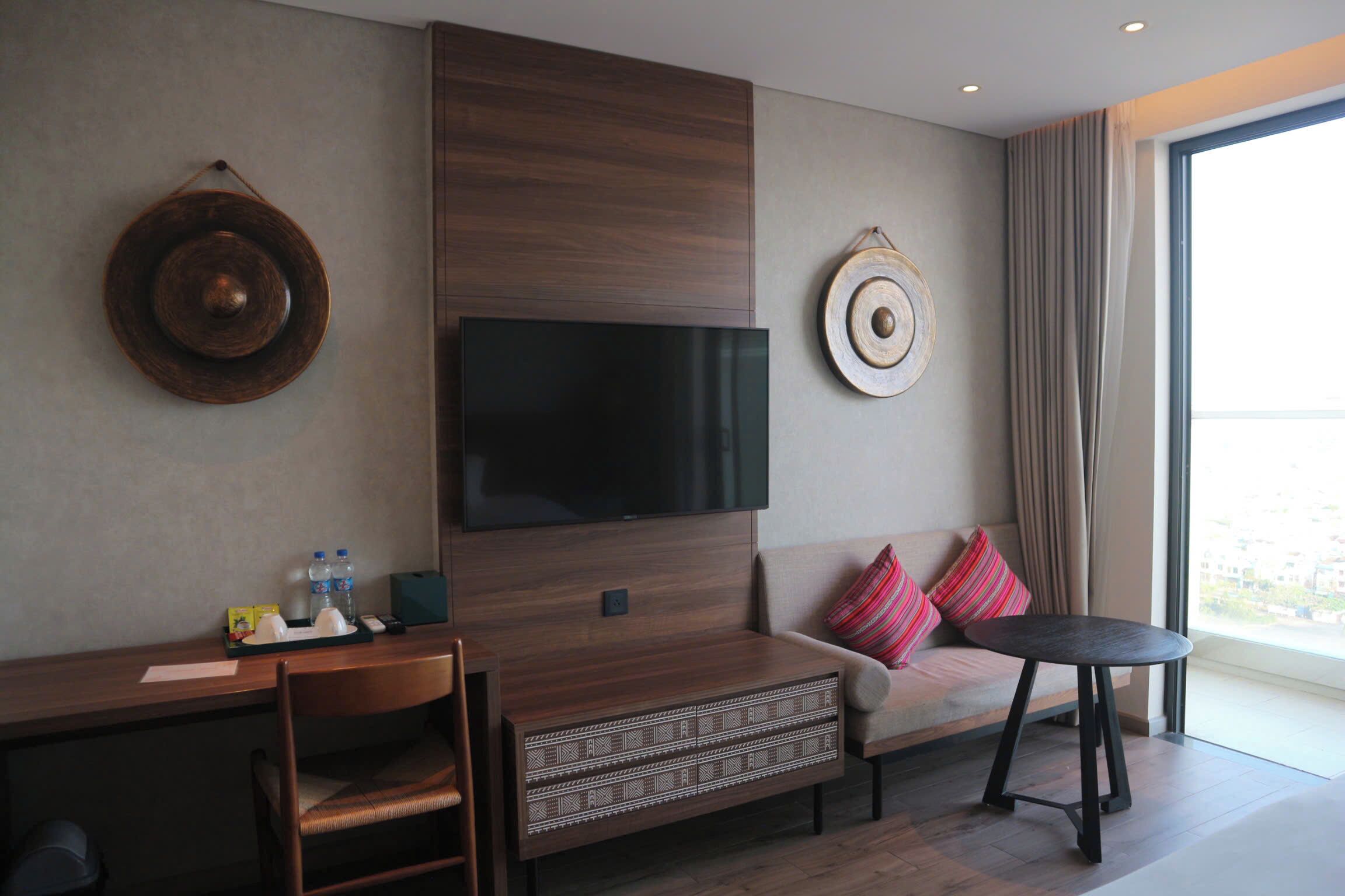 A modern hotel room with a flat-screen TV mounted on a wooden panel, a sofa with colorful cushions, a small round table, and a large window offering a city view.