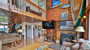 A spacious living room with a wooden floor, a fireplace, a television, and a high ceiling with wooden beams.