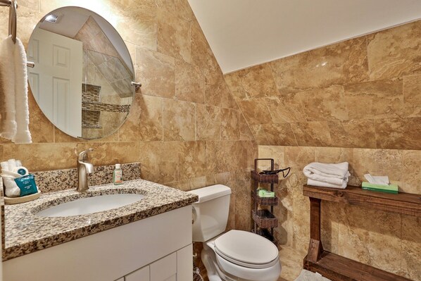 A bathroom with a granite countertop sink, a toilet, a mirror, and a wooden vanity with a basket.