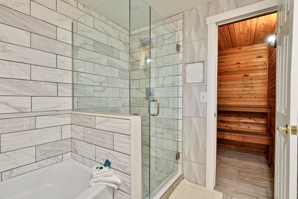 Un baño moderno con cabina de ducha de vidrio, una bañera blanca y una puerta de madera que da a una sauna.