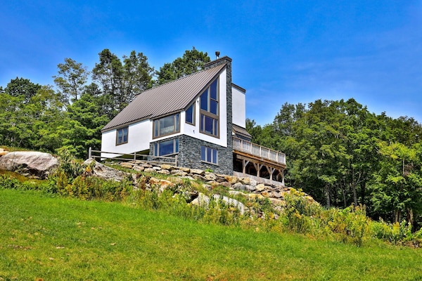 3-story Vermont chalet on sloping terrain, with lawn and trees all around beneath a clear blue sky.