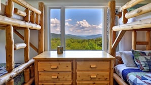 A cabin with a wooden interior, a large window offering a mountain view, and a wooden dresser with drawers.