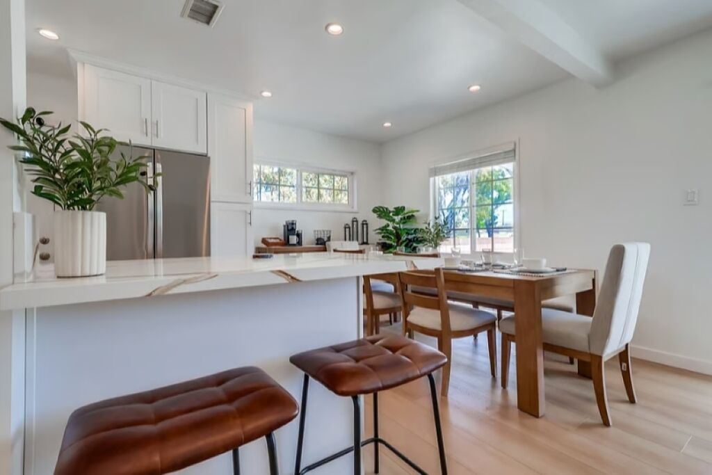 Une cuisine moderne avec des meubles blancs, un îlot central doté de tabourets et un coin repas avec une table et des chaises en bois.