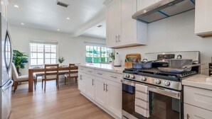 Una cocina moderna con armarios blancos, una estufa de acero inoxidable y una zona de comedor con mesa y sillas de madera.