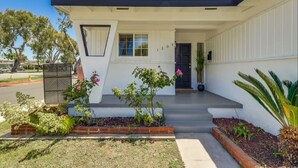 Una casa moderna de dos pisos con techo plano, un porche delantero y un pequeño jardín con diversas plantas y flores.