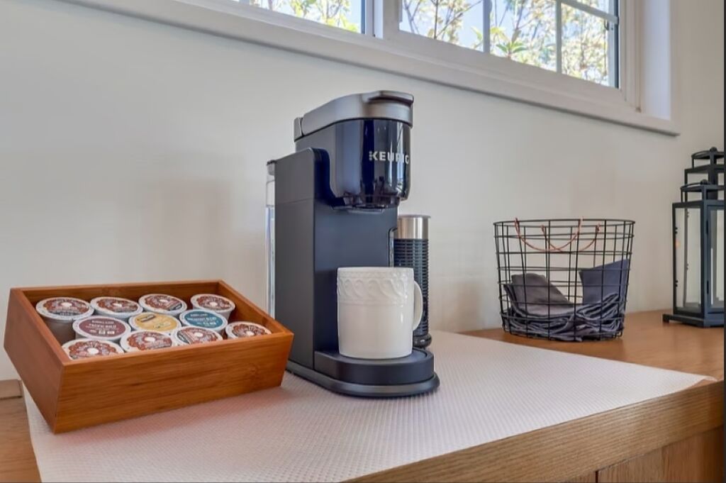 A coffee maker, a tray of coffee pods, a mug, and a basket on a table by a window.