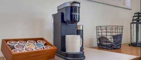A coffee maker, a tray of coffee pods, a mug, and a basket on a table by a window.