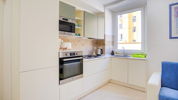 Una cocina compacta con electrodomésticos integrados, un ventanal con vistas a edificios y una silla azul.