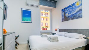 Un dormitorio con cama, una ventana con vistas a edificios, un cuadro en la pared y un marco con una fotografía.