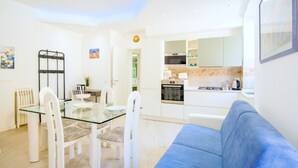 Una cocina moderna con armarios blancos, una zona de comedor con mesa de vidrio y sillas blancas, y un sofá azul en la sala de estar.