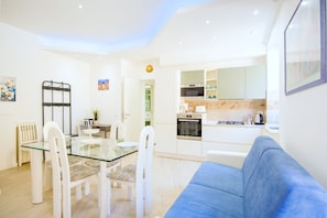 A modern kitchen with white cabinets, a dining area with a glass table and white chairs, and a blue sofa in the living area.