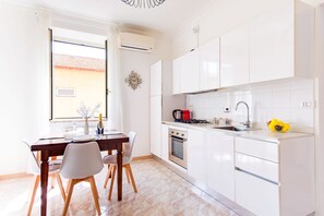 Uma cozinha moderna com armários brancos, uma mesa de jantar com cadeiras, forno, pia e janela com cortinas.
