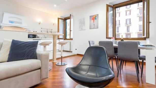 A modern living room with a sofa, a black chair, a dining area with a table and chairs, a kitchen area with a microwave, and a large window with a view of buildings.