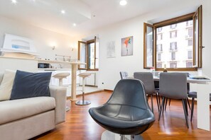 A modern living room with a sofa, a black chair, a dining area with a table and chairs, a kitchen area with a microwave, and a large window with a view of buildings. - Elegantis San Giovanni Suite - Chic Apartment with Bathtub Near Historic Cent (Roma)
