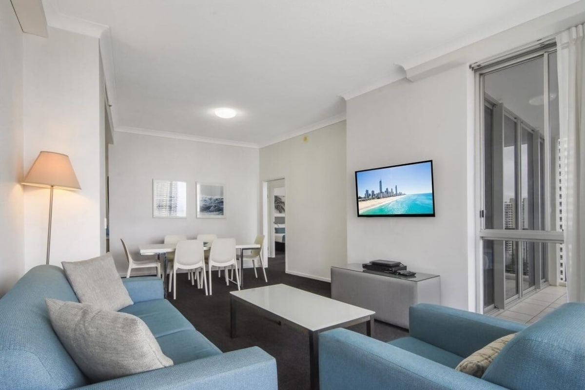 Uma sala de estar moderna com sofá, mesa de centro e TV exibindo uma vista urbana. Há uma área de jantar com cadeiras e mesa, e uma varanda com porta de vidro.
