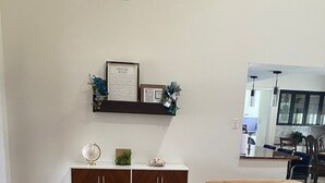 Un salón con suelo de madera, sofás beige, un mueble blanco con objetos decorativos y una cocina visible al fondo.