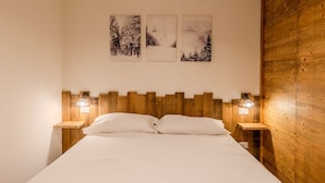 Une chambre à coucher avec une tête de lit en bois, un lit recouvert de draps blancs, deux tables de chevet avec des lampes et trois tableaux accrochés au mur représentant des paysages enneigés.
