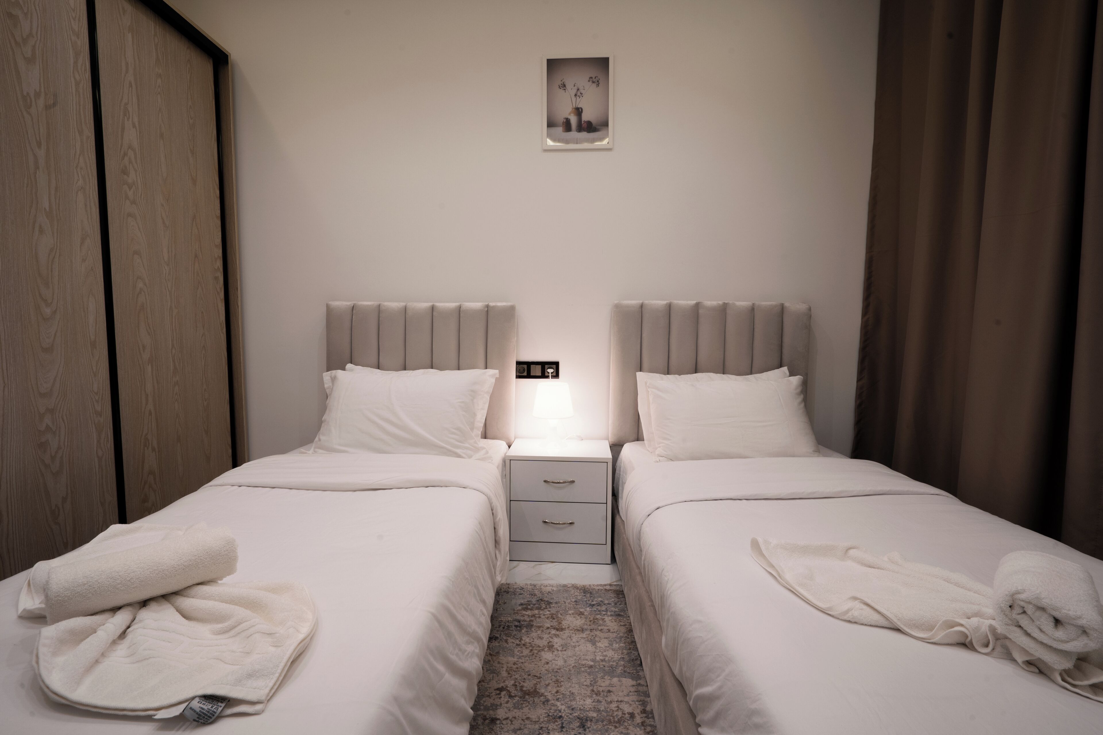 Una habitación de hotel con dos camas individuales, una mesita de noche, una pequeña foto en el marco en la pared, y una ventana con cortinas.
