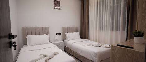 A hotel room with two beds, a bedside table, a nightstand, a potted plant, and a window with curtains.