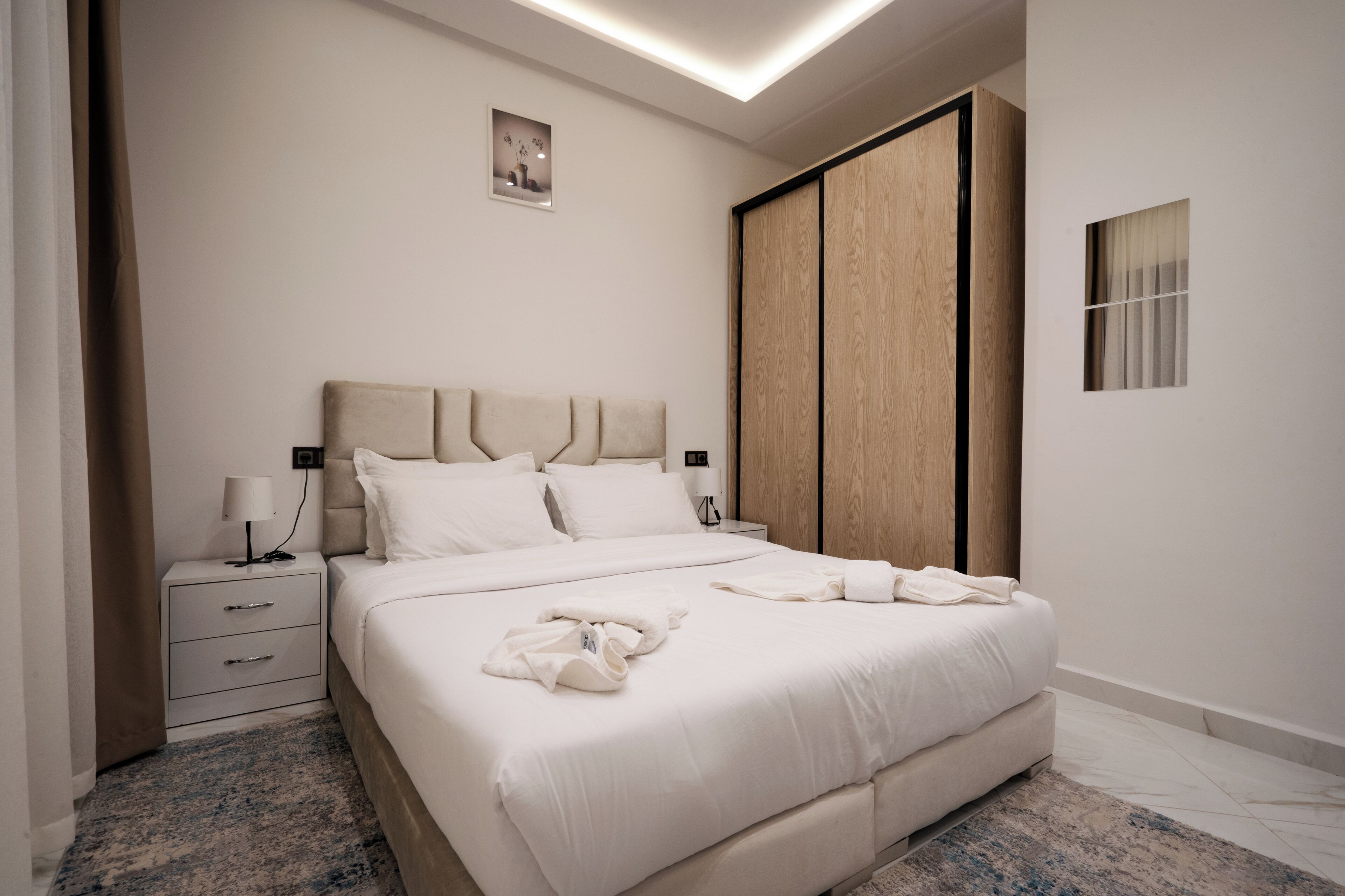 A modern bedroom with a large bed, a nightstand, a wardrobe, and a small window.