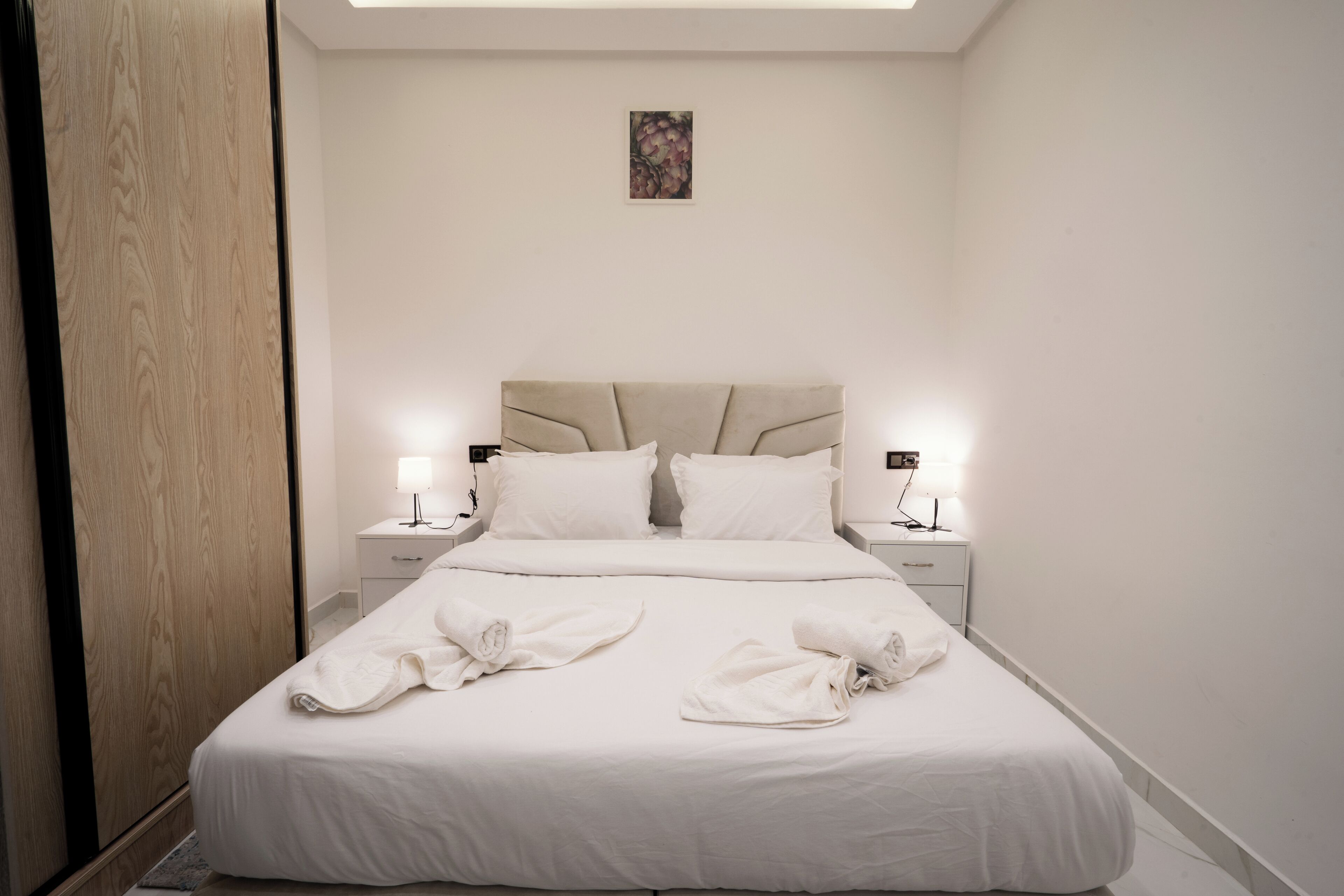 Una cama bien tendida con sábanas blancas y dos toallas dobladas encima, una mesita de noche con una lámpara, y un pequeño cuadro en la pared.