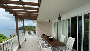 Ein überdachter Außenbereich mit Holztisch und weißen Stühlen, mit Blick auf ein Gewässer und viel Grün.