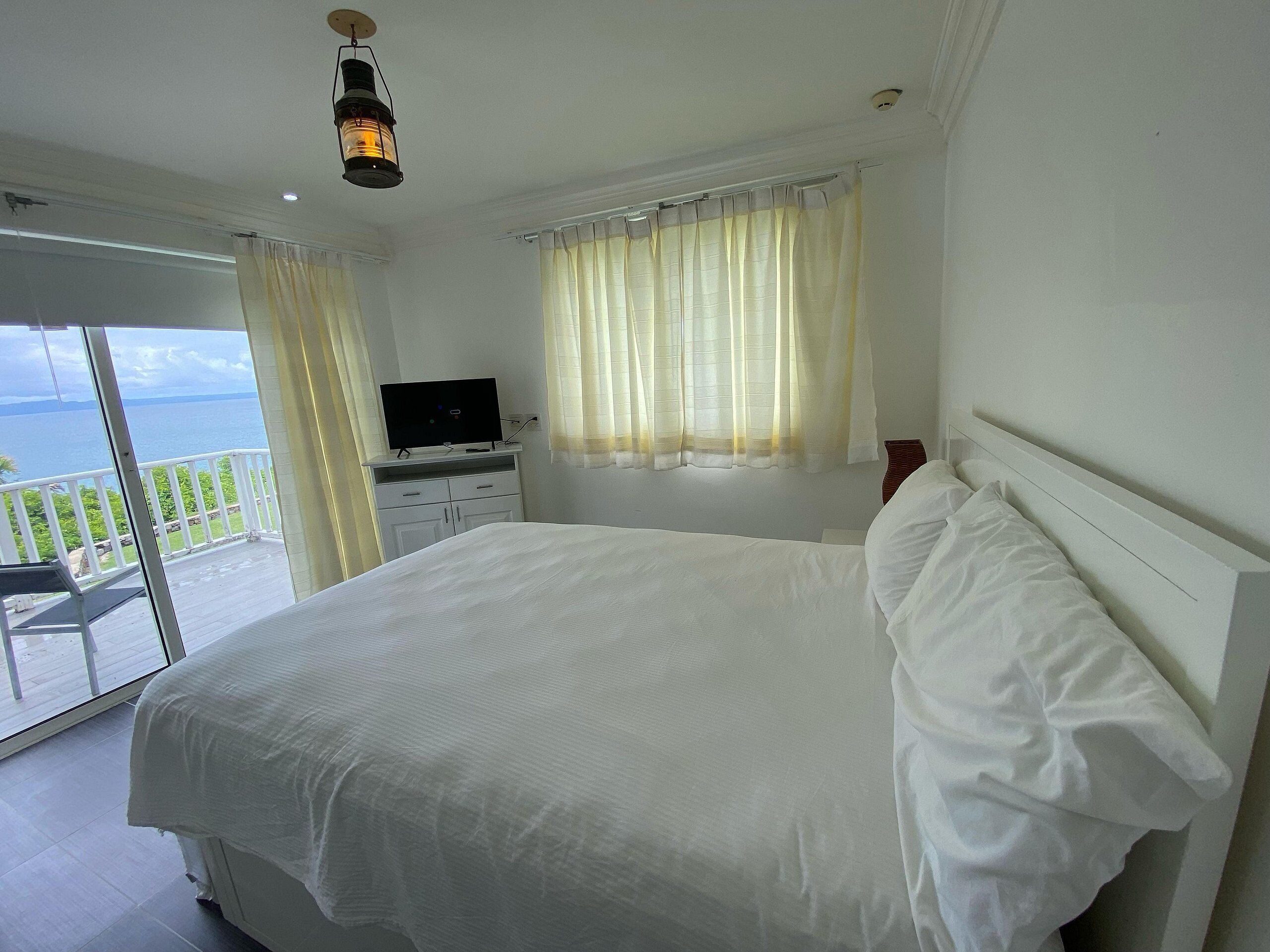 A bedroom with a large bed, a television on a dresser, and a balcony with a view of the sea.