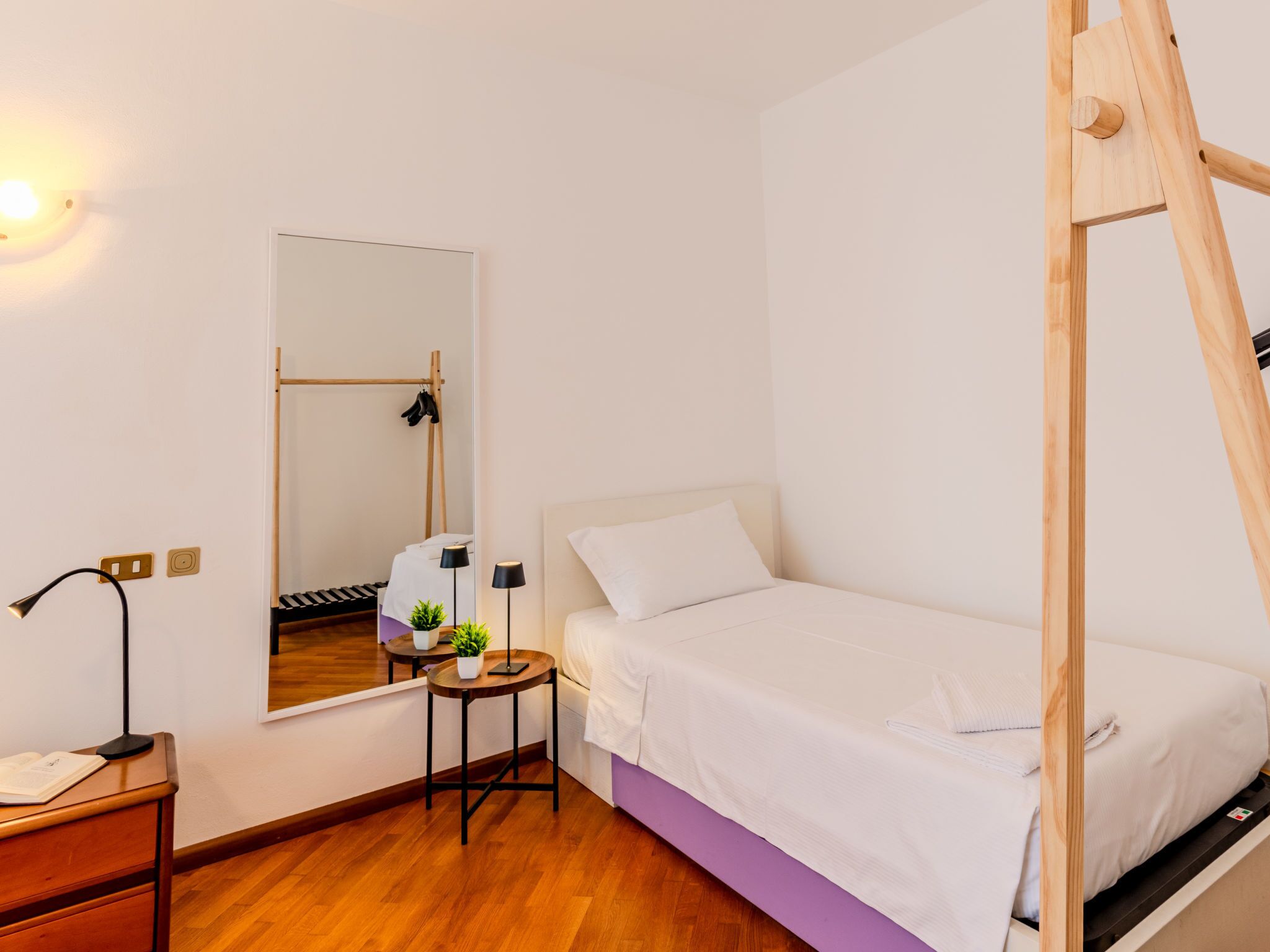 Une chambre à coucher avec un lit, une échelle en bois, un miroir, un bureau avec une lampe et une petite table avec une plante.