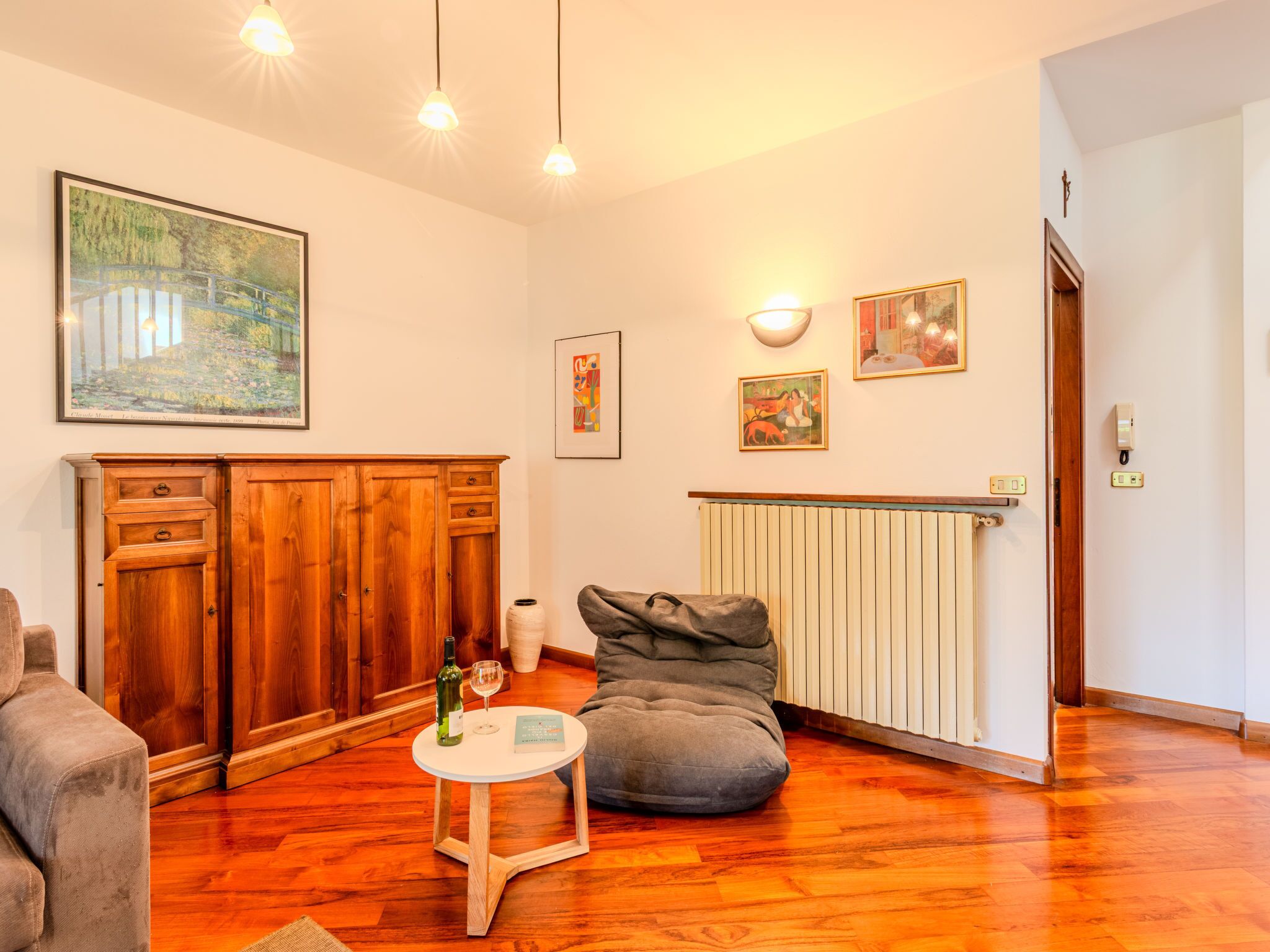 A cozy living room with wooden flooring, a grey bean bag, a small round table with a wine bottle and glass, and a painting on the wall.