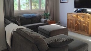 A living room with a grey sectional sofa, a wooden cabinet with a flat-screen TV, and a large window with curtains. - Charming 3-bedroom house walking distance to the wonderful Napier, marine parade (Napier)