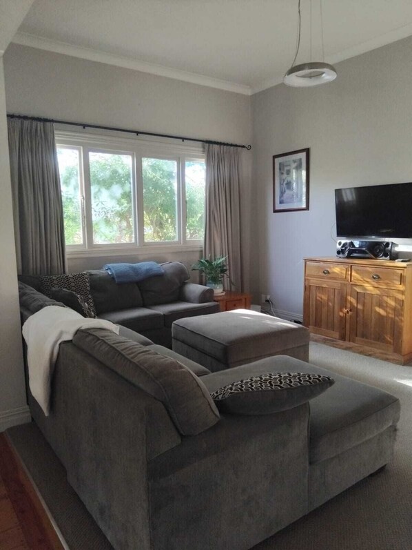 A living room with a grey sectional sofa, a wooden cabinet with a flat-screen TV, and a large window with curtains. - Charming 3-bedroom house walking distance to the wonderful Napier, marine parade (Napier)