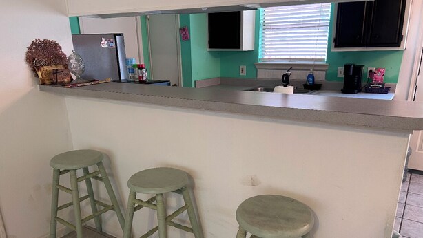 A kitchen with a white countertop, three green stools, a refrigerator, and a window with blinds.