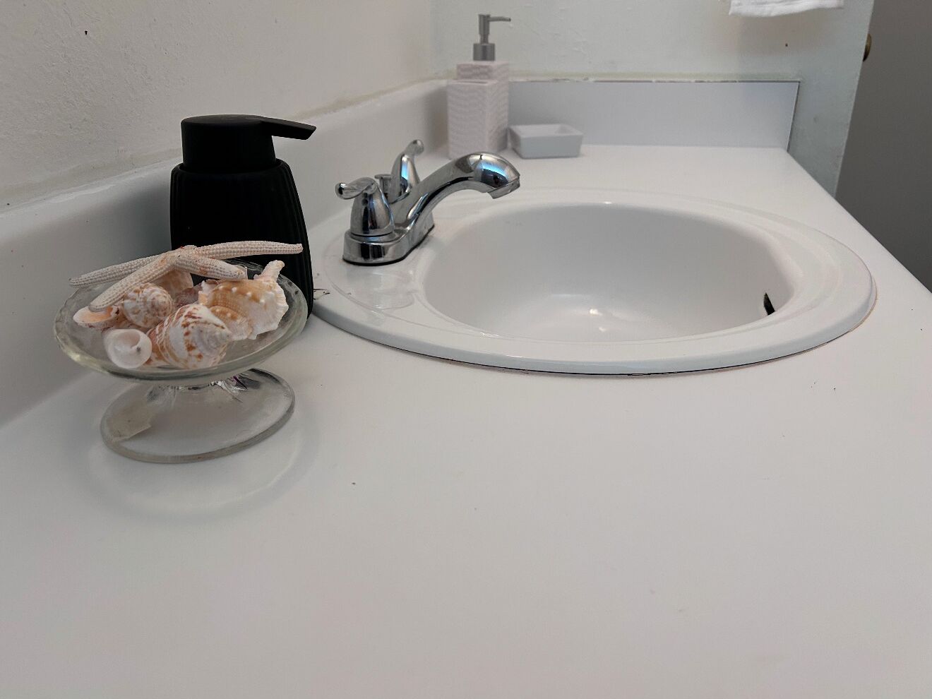Ein Badezimmerwaschbecken mit chromfarbenem Wasserhahn, eine Glasvase mit Meeresschnecken und ein schwarzer Seifenspender.