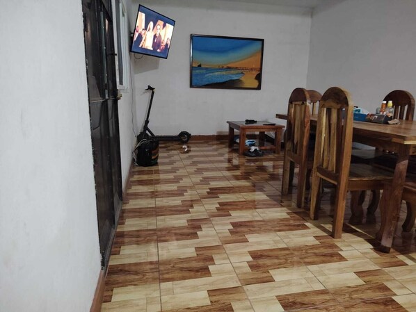 Ein Zimmer mit gefliestem Boden, einem an der Wand befestigten Fernseher, einem Gemälde und einem Esstisch mit Stühlen.