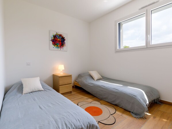 A small, bright room with two single beds, a nightstand with a lamp, a colorful abstract painting, and a large window.