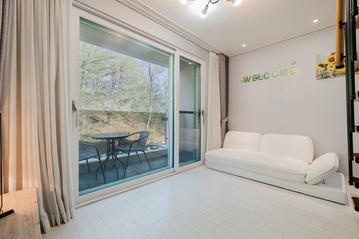 Un moderno salón con un sofá blanco, un ventanal de vidrio que da a una zona de estar exterior, y un cartel con la palabra 'WELCOME' en la pared.