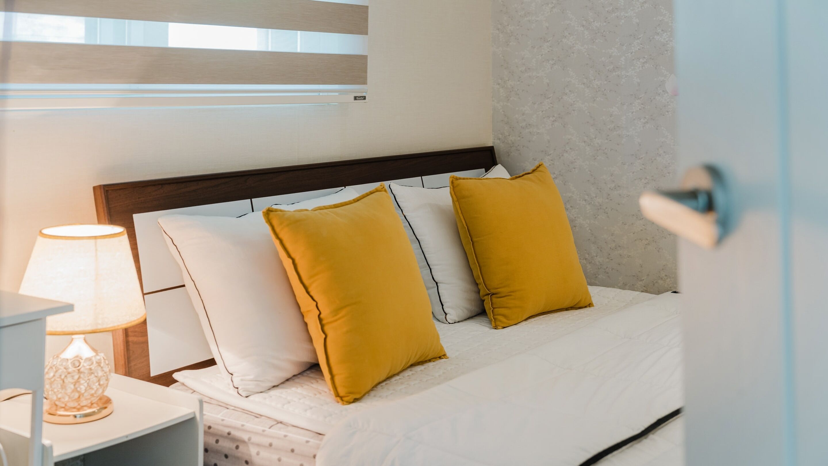 Un lit bien fait, avec des oreillers blancs et jaune moutarde, une lampe de chevet et une tête de lit en bois.