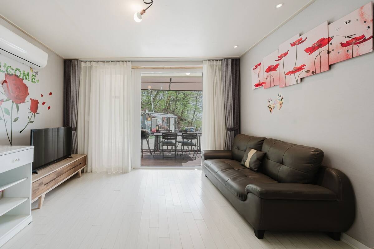 Una sala moderna con un televisor de pantalla plana, un sofá de cuero y un comedor visible a través de una puerta de cristal.