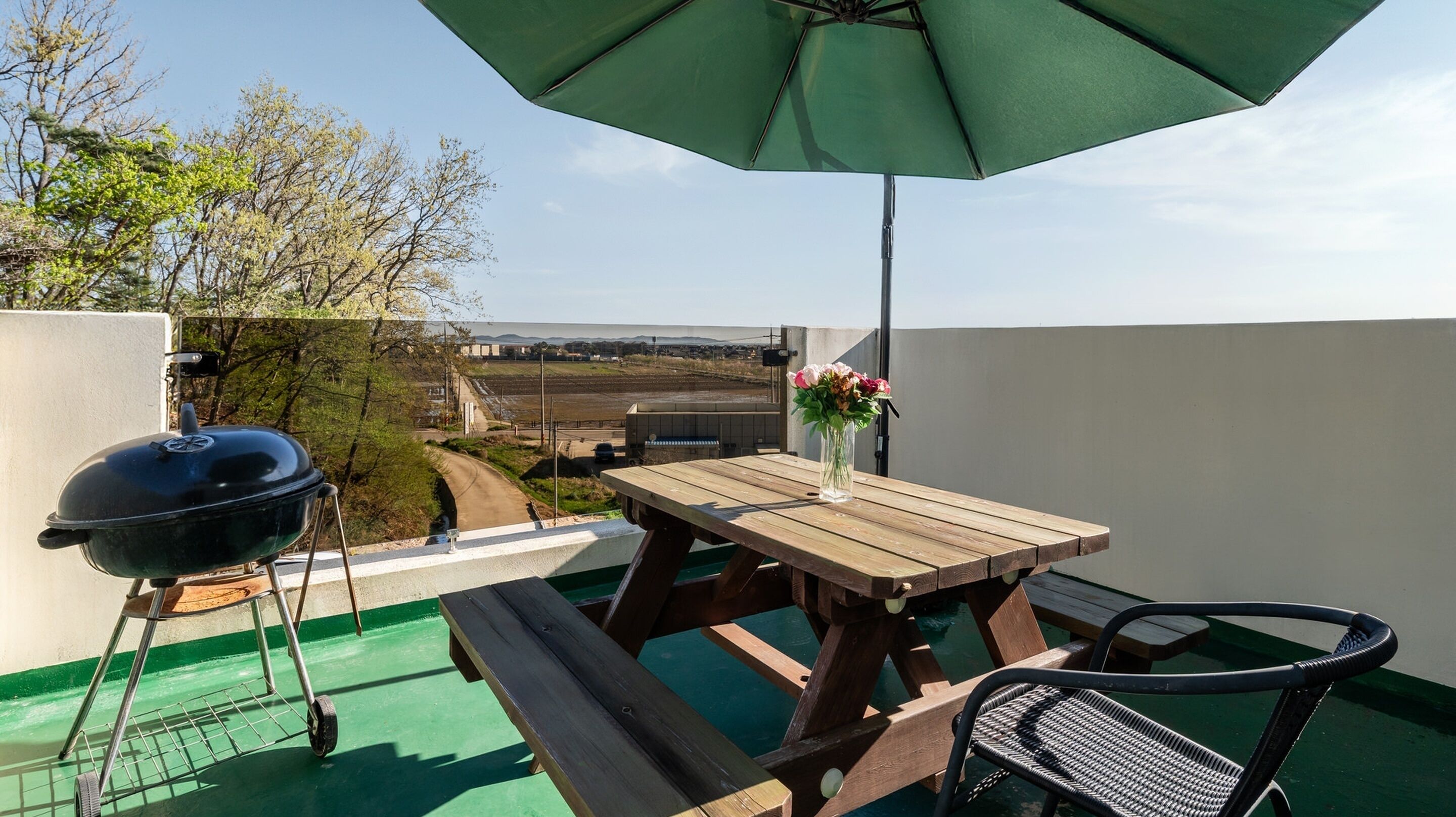 A wooden table with a green umbrella, a barbecue grill, and a wicker chair on a balcony with a view of a road and buildings.
