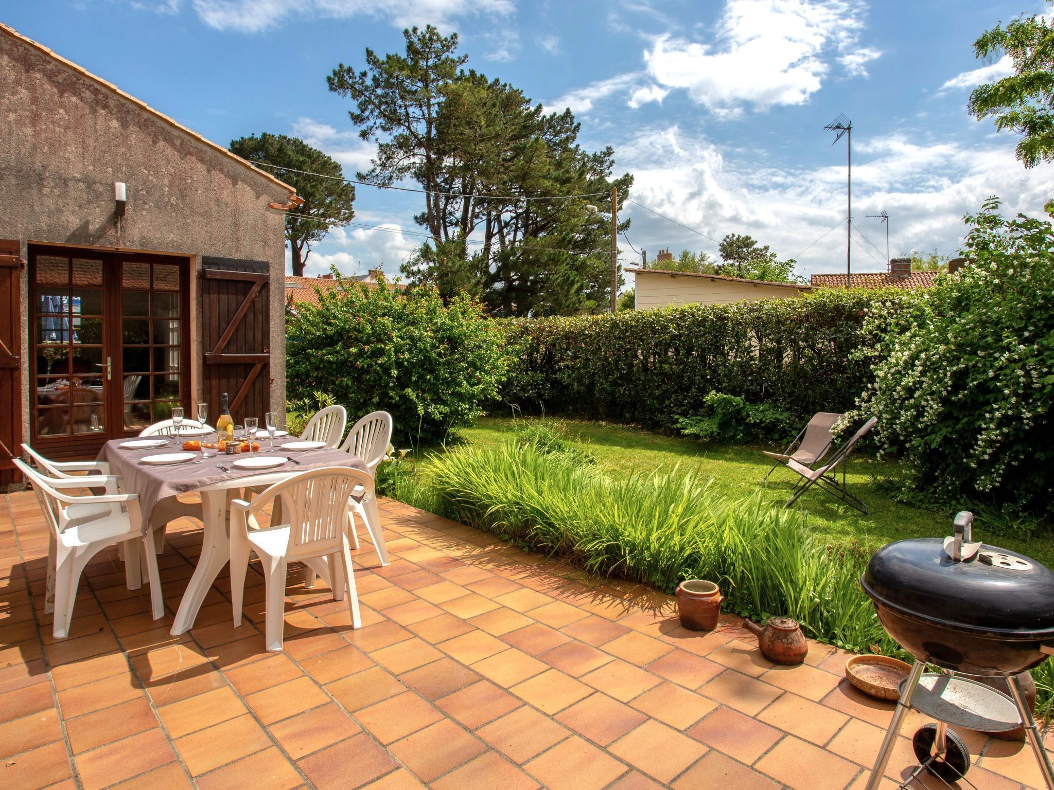 Um pátio com mesa posta para refeições, cercado por um jardim com churrasqueira e um sebe.