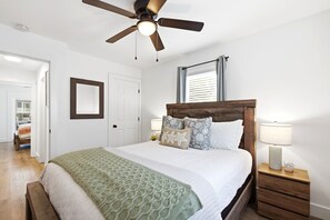 A bedroom with a bed, a ceiling fan, a nightstand with a lamp, and a window with blinds.