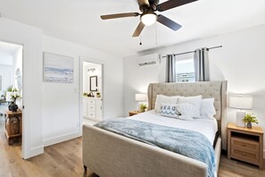 Un dormitorio con cama, mesitas de noche, ventilador de techo, ventana con cortinas y aire acondicionado de pared.