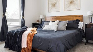 Un dormitorio con una cama grande, ropa de cama oscura, dos cuadros en la pared, una mesita de noche con lámpara y una ventana con cortinas.