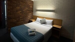 A hotel room with a brick accent wall, a white brick wall, a bed with a blue patterned bedspread, two pillows, and a bedside table with a lamp. - Arabella Motor Inn (Tweed Heads South)