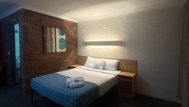 A hotel room with a bed, a nightstand, a wall-mounted light, and a brick accent wall. - Arabella Motor Inn (Tweed Heads South)