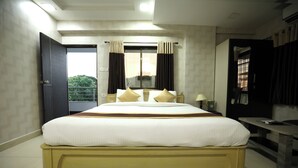 A large bed with a wooden frame and white bedding, a bedside table with a lamp, and a view of the outdoors through a window with curtains. - Hotel Park Gardenia (Nagpur)