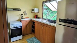 A compact kitchen with a stove, microwave, and refrigerator, wooden cabinets, and a window with a view of greenery.