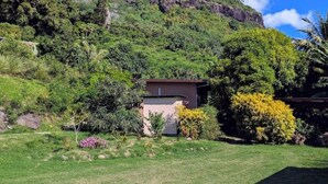 A house with a small garden, surrounded by lush greenery and a hill in the background.