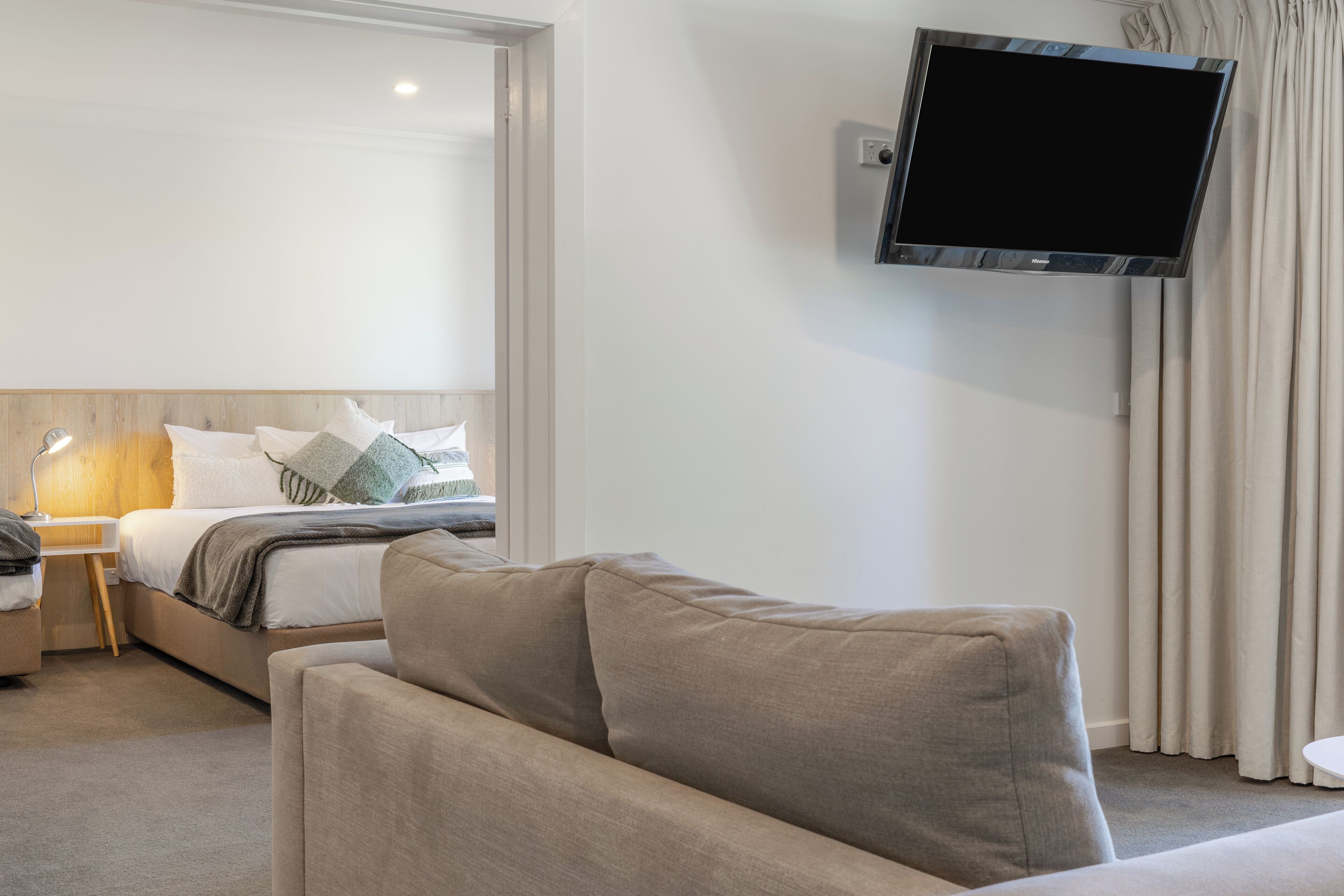 A modern hotel room with a bed, a sofa, a flat-screen TV mounted on the wall, and a bedside table with a lamp.